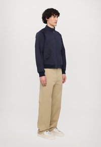 Giubbotto bomber blu navy con colletto alto, zip davanti, tasche laterali e polsini a coste, abbinato a pantaloni beige a gamba larga e sneakers bianche.