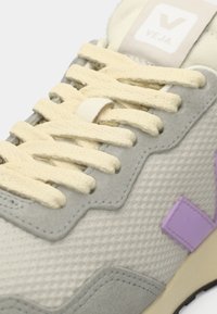 Sapatilha cinza claro com um cabedal texturizado, atacadores creme e um detalhe roxo de lado. Apresenta uma sola de borracha e uma língua com marca.