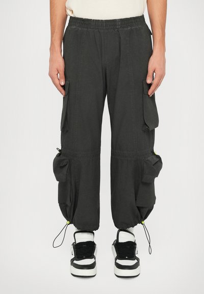 Μαύρο cargo παντελόνι με ελαστική μέση, πολλές πλαϊνές τσέπες, ρυθμιζόμενους μανίκια και λεία υφή, συνδυασμένο με μαύρα και άσπρα αθλητικά παπούτσια.