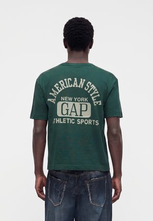 Osoba otočená chrbtom, má na sebe zelené tričko s nápisom „American Style New York Gap Athletic Sports“ na chrbte a modré džínsy.