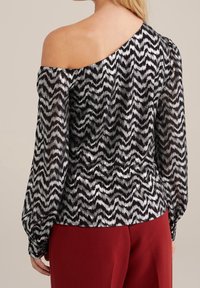 Černá a stříbrná halenka s odhalenými rameny se zigzagovým vzorem, dlouhými nadýchanými rukávy a průsvitnou texturou, doplněná červenými kalhotami.