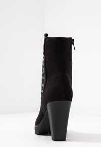 Botín de ante negro con tacón grueso, que presenta una cremallera lateral y un detalle de logo prominente a lo largo del costado. Textura suave y diseño elegante.