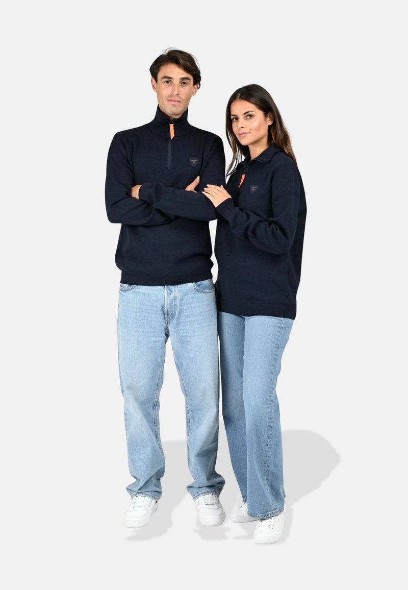Pulls en laine zippés navy en tissu doux, avec des accents de fermeture éclair orange contrastants. Associés à un jean bleu clair et des baskets blanches.