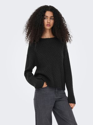 Jeune femme aux longs cheveux bouclés portant un pull noir texturé et un jean gris foncé, debout avec une main dans sa poche.