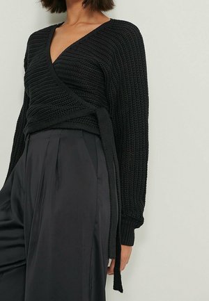 Femme portant un pull en maille noire cache-cœur à manches longues et un pantalon noir plissé taille haute sur un fond uni.