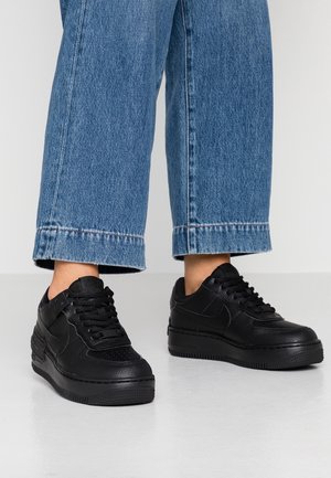 Personne portant un jean bleu droit et des baskets Nike Air Force 1 noires sur un sol blanc avec un fond clair uni.