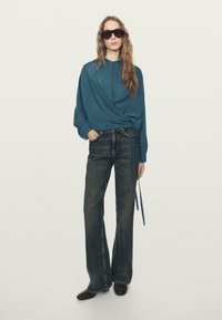 Bluse i blågrøn farve med lange ærmer og bindedetalje, kombineret med højttaljede mørke denim flare jeans og brune sko. Modellen bærer sorte solbriller.