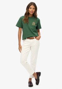 T-shirt en coton vert avec un logo doré, pantalon blanc à jambe droite et mocassins bordeaux foncé avec une semelle texturée. Coupe décontractée et style relaxé.