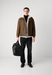 Brun pufferjakke med en cremefarvet faux fur krave, beige cardigan underneath, sorte jeans og en sort rejsetaske. Sorte sko fuldender looket.