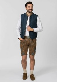 Traditionelles bayerisches Outfit mit einer dunkelblauen Steppweste, weißem Hemd, braunen Lederhosen mit blauer Blumenstickerei und braunen Schuhen.
