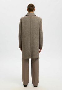 Cappotto marrone a motivo houndstooth con colletto a punta, maniche lunghe e silhouette dritta. Abbinato a pantaloni palazzo beige chiaro.