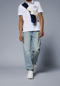 Osoba ubrana w białą koszulkę polo Tommy Hilfiger, jasnoniebieskie dżinsy, białe trampki oraz granatowo-białą w paski bluzę narzuconą na ramię, idąca do przodu.