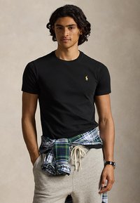 Ung mand med krøllet hår iført sort kortærmet skjorte med gul logo, grå joggingbukser, ternet skjorte bundet om livet og et ur.