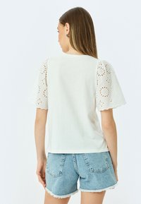 Hvid skjorte med bølget eyelet-detalje på ærmerne, parret med lyseblå denimshorts. Blødt stof, afslappet pasform og casual design.