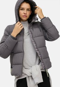 Grå pufferjakke med hætte, lynlåslukning og ribbede manchetter. Designet er quiltet med en blank finish. Modellen bærer en grå skjorte under.