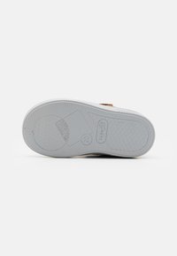 Semelle de chaussure en caoutchouc blanc avec un motif de grip texturé, comportant des nervures et un détail circulaire. Dessus marron avec un hardware de sangle au-dessus de la semelle.