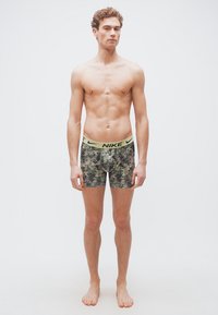 Νέος άνδρας στέκεται ξυπόλυτος φορώντας μποξέρ Nike με σχήμα καμουφλάζ και ανοιχτοπράσινη μέση, μπροστά σε απλό φόντο.