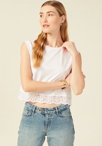 Witte katoenen crop top met kantafwerking aan de zoom, gecombineerd met lichtblauwe high-waisted jeans. Het model heeft lang bruin haar en draagt een gouden armband.