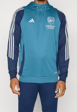 Person iført blå og navy Arsenal fodbold-hættetrøje og bukser med Adidas-striber og Emirates Fly Better-logo på brystet.