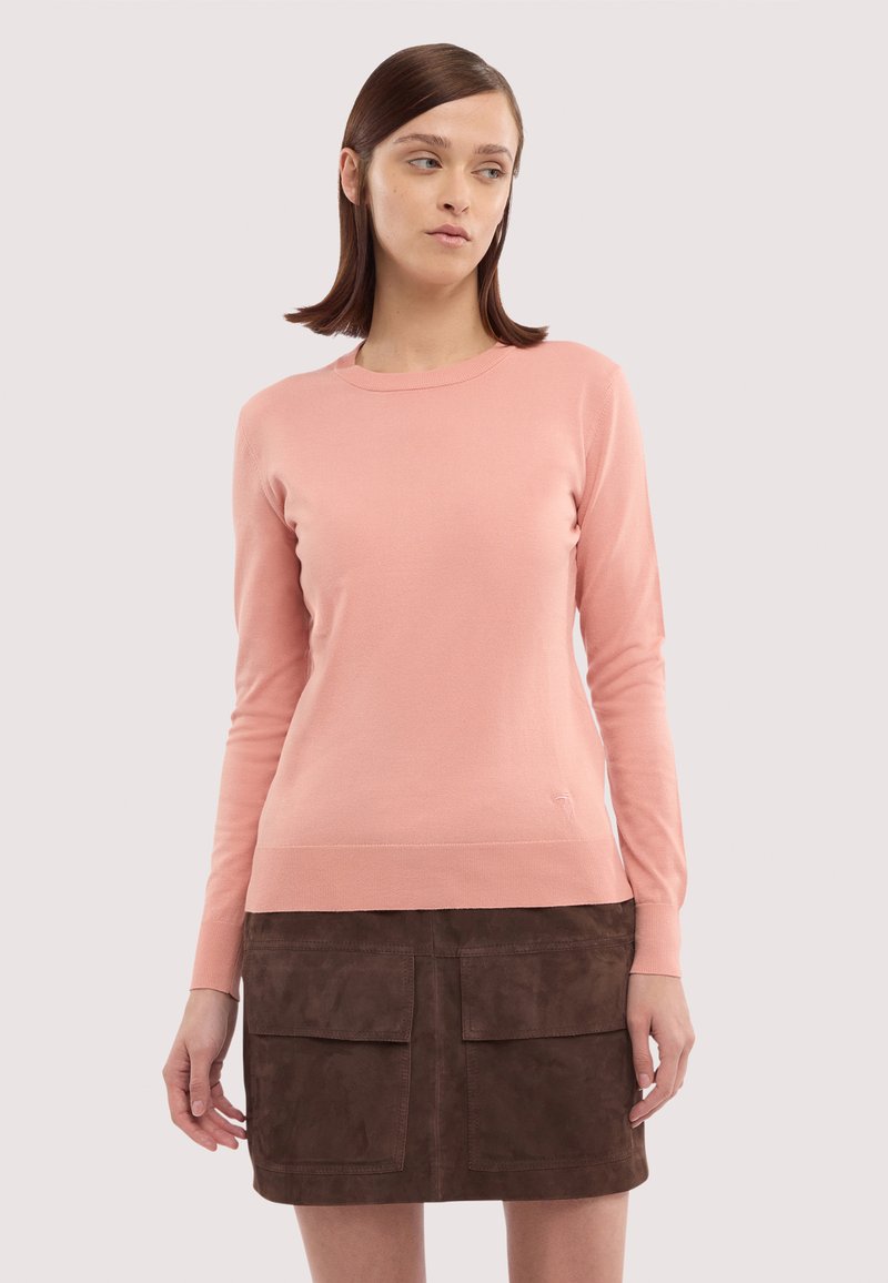 Frau trägt einen hellrosa Pullover mit langen Ärmeln und einen dunkelbraunen Rock mit Vorderentaschen, steht vor einem neutralen Hintergrund.