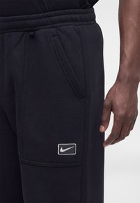 Άνδρας με μαύρη φόρμα Nike που δείχνει την πλαϊνή τσέπη και την ετικέτα με το λογότυπο, με το δεξί χέρι χαλαρό στο πλάι, σε φωτεινό φόντο.