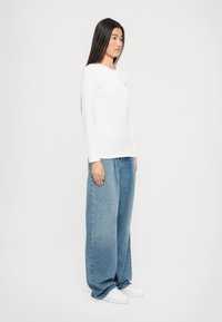 Hvit langermet topp med rund hals, kombinert med løstsittende blå denim jeans. Modellen har på seg hvite joggesko og står foran en hvit bakgrunn.