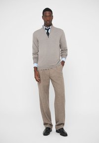 Suéter bege claro com decote em V, combinado com calças de tweed castanhas. Sapatos pretos completam o visual. Fundo simples.