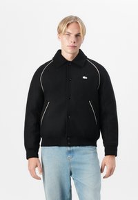 Jachetă din fleece negru cu un design cu guler, având accente albe, nasturi pe partea din față și un logou mic pe piept.