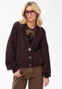 Brązowy sweter z dzianiny z wzorem w romby, z głębokim dekoltem w kszta�łcie litery V i trzema dużymi guzikami. Nosi się go na przezroczystą bluzkę w kwiaty i w parze z brązowymi spodniami.