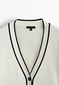 Cardigan beige clair avec un col en V profond, bordure noire, et une fermeture à un seul bouton. La texture est tricotée et lisse, avec un motif subtil.