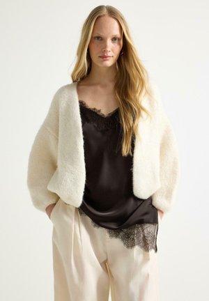 Kvinde med langt blondt hår iført en cremefarvet, pelsagtig cardigan, sort silketop med blonder samt løse cremefarvede bukser, stående mod en hvid baggrund.