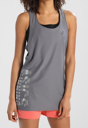 Frau trägt ein graues ärmelloses Asics Running-Top über einem schwarzen Sport-BH und korallenfarbene Sportshorts, steht mit entspannten Armen.