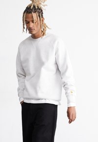 Grå crewneck sweatshirt med ribbede manchetter og kant, med et lille gult logo på venstre ærme. Båret over en hvid skjorte.