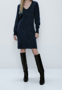 Robe tricotée à encolure en V profonde, de couleur bleu marine foncé, avec des manches longues ; associe des bottes en cuir noir hautes ; texture lisse et coupe décontractée.