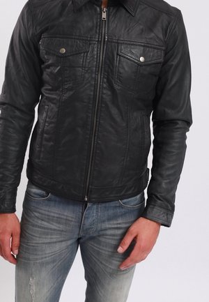 Zwarte leren jas met een ritssluiting aan de voorkant, twee borstzakken en een gestructureerde kraag. Draagt over lichtblauwe versleten spijkerbroek.