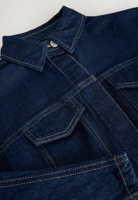 Denimjacka i mörkblått med klassisk krage, två bröstfickor och kontrasterande orange sömmar. Slät textur och strukturerad form.
