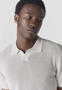 Jeune homme aux cheveux courts et bouclés regardant vers le bas, portant un polo blanc en maille ainsi que des piercings au nez et aux oreilles, sur un fond uni.