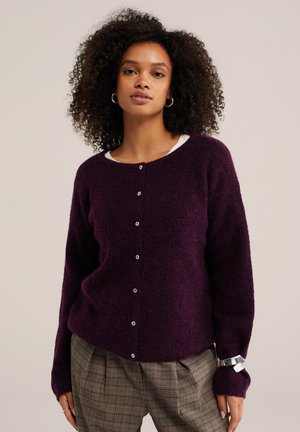Frau mit lockigem Haar, die einen lila Knopf-Pullover und karierte Hosen trägt und vor einem schlichten beigen Hintergrund steht.