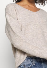 Hellbeiger gestrickter Pullover mit V-Ausschnitt und geripptem Muster. Lange Ärmel und lockere Passform, kombiniert mit blauen Jeans.