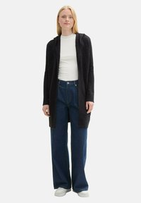 Lång, mörk stickad cardigan med huood, över en vit ribbad topp, kombinerad med lösa blå denimjeans och vita sneakers.