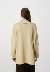 Ribbet beige genser med høy krage, lange ermer og et merke på baksiden. Mykt, teksturert stoff med løs passform og sideslisser.