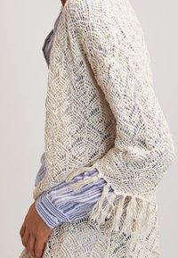 Gebreide sjaal met een diamanten patroon in crème, met franjes aan de randen. Draagt over een blauw-wit gestreept shirt, wat het open weefontwerp benadrukt.