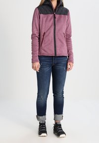 Polarowa kurtka z czarną pikowaną górą, z zamkiem z przodu i dwoma bocznymi kieszeniami. Kolor wina o gładkiej teksturze. Połączona z niebieskimi dżinsami.