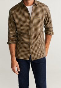 Hnědočerná kostkovaná košile na knoflíky z bavlny, s klasickým límcem, dlouhými rukávy a přední kapsou. Doplněna tmavými džínami.