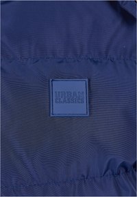 Giacca trapuntata blu navy in tessuto dalla superficie liscia. Presenta un'etichetta con logo quadrato in rilievo etichettata "URBAN CLASSICS" al centro.