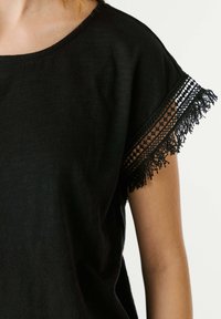 Black short-sleeve top with lace and fringe detailing on sleeve edge, worn on arm and shoulder against a plain background.