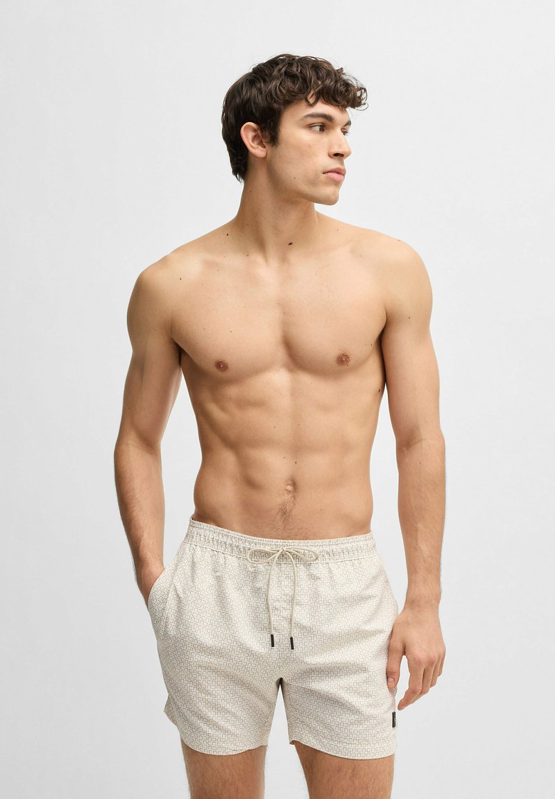 Beige Badehose mit geometrischem Muster, elastischem Bund, Kordelzug und seitlichen Taschen, getragen von einem oberkörperfreien männlichen Modell.