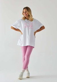 Luźna biała koszulka z różowym napisem graficznym, w zestawie z miękkimi różowymi legginsami i białymi trampkami z różowymi i zielonym akcentami.
