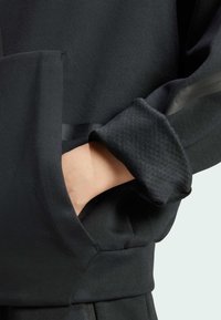 Crni atletski pulover s teksturiranim manžetama i kontrastnim glatkim obrubom. Ima džep i vidljivo šivanje uz šavove.