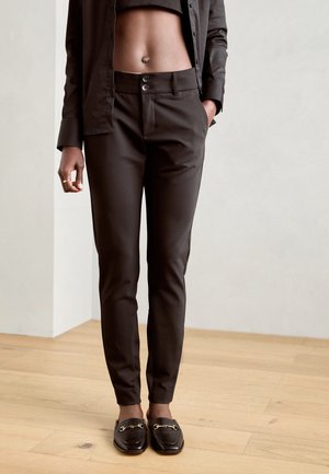 Pantalons ajustés marron foncé avec une coupe slim, dotés de deux boutons à l'avant, de passants de ceinture et de poches latérales, associés à des mocassins noirs.